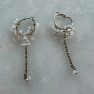Silvriga örhängen med vita pärlor och kedja - Unika silvriga örhängen med små vita pärlor och dekorativ kedja. Örhängena har en rund form och pärlorna är fästa med guldfärgade detaljer. Perfekta för dig som vill ha något annorlunda och stilrent.