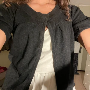 Svart kofta med kort ärm - Säljer en svart kofta från Vero moda med korta ärmar och rund halsringning. Koftan har en enkel design med en knapp upptill. Perfekt att slänga över en topp eller klänning. Saknar en knapp på ena sidan, men annars i gott skick! 