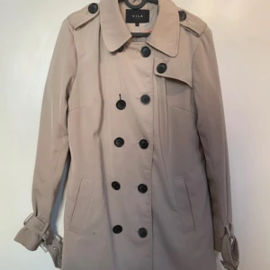 Beige trenchcoat från Vila - Stilren beige trenchcoat från Vila med dubbelknäppning och klassisk krage. Jackan har långa ärmar med spännen vid handlederna och två fickor framtill. Perfekt för övergångssäsonger och ger en tidlös look.
