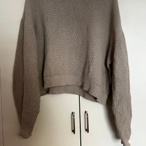Beige stickad croppad tröja - Säljer en beige stickad croppad tröja med lång ärm och avslappnad passform. Perfekt att slänga på sig över ett linne eller t-shirt. Enkel och stilren design som passar till det mesta.