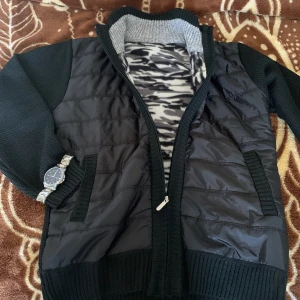 Svart stickad och quiltad jacka med dragkedja - Säljer en svart cardigan med stickade ärmar och rygg samt quiltad framsida. Cardiganen har dragkedja och ribbade muddar. Insidan har ett mönstrat foder i grått och svart. Perfekt för blåsiga dagar. Du ansvar för frakt.