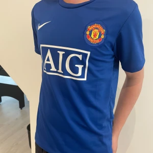 Manchester United blå fotbollströja Nike - Blå Manchester United bortatröja med vitt AIG-tryck på bröstet och klubbmärke på vänster sida. Klassisk Nike-logga i vitt. Rund hals och korta ärmar. Perfekt för fotbollsfans som vill sticka ut på planen eller läktaren.