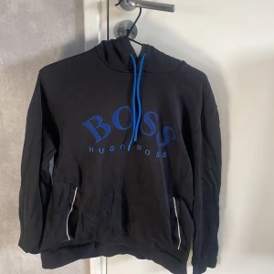 Svart hoodie från Hugo Boss med blå detaljer - Säljer en svart hoodie från Hugo Boss med blå snören och blått BOSS-tryck på bröstet. Tröjan har huva och fickor med vita detaljer.