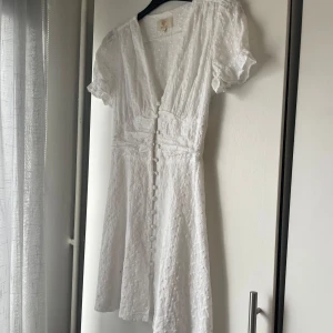 Vit kortklänning med broderade detaljer och knappar - Superfin vit kortklänning med broderade blommor, v-ringning och puffärmar. Klänningen har knappar framtill och markerad midja med smock baktill. Perfekt för sommarens alla tillfällen! Den sista bilden är hur den ser ut på mig och den är lite för liten i bysten för mig så den ska inte se ut så där🥰❤️