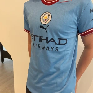 Manchester City fotbollströja 2022/23 - Säljer en ljusblå Manchester City hemmatröja från Puma med klubbmärke och sponsortryck på bröstet. Tröjan har röda och vita detaljer vid krage och ärmslut. Perfekt för dig som vill visa ditt stöd på matchdag!