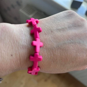 Rosa armband med kors - Säljer ett coolt armband i starkt rosa färg med flera korsformade pärlor. Perfekt accessoar för att addera färg till din stil. Stretchigt och bekvämt att bära.