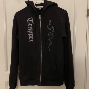 Svart hoodie med strasstenar från Trapper - Svart hoodie från Trapper med dragkedja, huva och fickor. Dekorerad med strasstenar i form av en orm och texten 'Trapper' på framsidan. Perfekt för en cool och avslappnad stil.