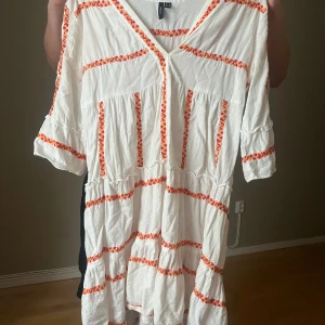 Vit kortklänning med orange broderi - Superfin vit kortklänning från H&M med orange broderade ränder och v-ringning. Klänningen har trekvartsärm och är gjord i luftig bomull, perfekt för varma dagar. Snyggt avslappnad passform och volangdetaljer för en bohemisk vibe.