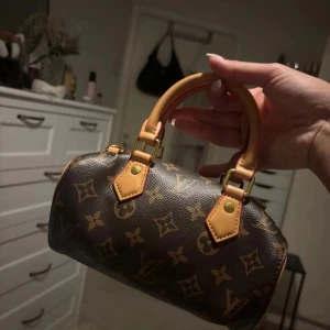  handväska med långt band - Säljer en ikonisk liten handväska med klassiskt monogrammönster i brunt och beige. Väskan har hjärt form, två korta handtag i ljust skinn och guldfärgade metalldetaljer. Dragkedja upptill och snygga detaljer som gör den superchic.
