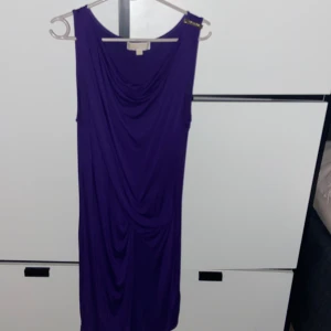 Lila långklänning Michael Kors S - Snygg lila långklänning från Michael Kors i mjukt stretchigt material. Klänningen är ärmlös med draperad front och en liten gulddetalj på axeln. Perfekt för dig som vill sticka ut med en elegant och stilren look. Den är endast testad