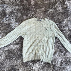 Grå kabelstickad tröja Polo Ralph Lauren - Kabelstickad tröja från Polo Ralph Lauren i ljusgrått med klassisk rund halsringning och broderad logga på bröstet. Tröjan är långärmad och har ribbade muddar vid ärmslut och nederkant. Perfekt för lager-på-lager och snygg till jeans.