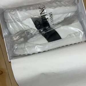 Valentino Garavani vita sneakers - Snygga vita sneakers från Valentino Garavani med svart band över vristen och logga på sidan. Skorna har en chunky sula med nitar och är tillverkade i skinn. Perfekta för dig som vill ha en clean och lyxig look med streetkänsla.