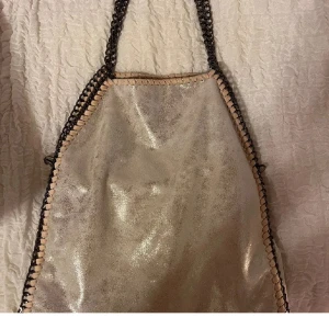 Glittrig silver axelväska med kedja - Snygg axelväska i glittrigt silver med kedjedetaljer längs kanterna och som axelrem. Väskan har mjukt material, ljusbeige kant och dragkedja på baksidan. Rymligt innerfack med extra fickor för småsaker. Perfekt för dig som vill sticka ut.