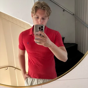 Röd pikétröja från Ralph Lauren - Säljer en klassisk röd pikétröja från Ralph Lauren med blå broderad logga på bröstet. Tröjan har korta ärmar, krage och knappar framtill. Perfekt för en clean och sportig look. Tillverkad i mjuk bomull som andas. Sitter som Xs/S
