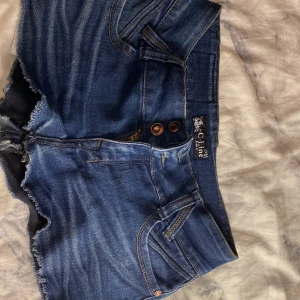 Blå jeansshorts från C-Line - Snygga blå jeansshorts från C-Line med råa kanter och dubbla knappar framtill. Klassisk femficksmodell med hjärtformade bakfickor och slitna detaljer för en cool look. Perfekta för varma dagar och enkel att matcha med allt.