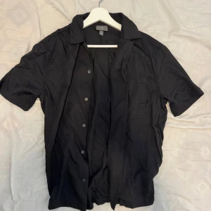 Tre kortärmade skjortor i olika färger - Säljer tre stilrena kortärmade skjortor. En svart, en marinblå och en beige skjorta, alla med klassisk krage och knäppning framtill. Perfekta för en clean och avslappnad look. Skjortorna är i mjukt material och har en normal passform.