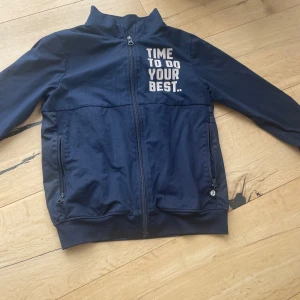 Marinblå vindjacka från Pomp de Lux - Snygg marinblå vindjacka från Pomp de Lux med trycket 'TIME TO DO YOUR BEST.' på bröstet. Jackan har hel dragkedja framtill, två fickor med dragkedja och ribbade muddar. Perfekt för dig som gillar sportig och clean stil.