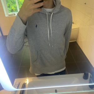 Grå hoodie från Polo Ralph Lauren - Snygg grå hoodie från Polo Ralph Lauren med klassisk broderad logga på bröstet. Tröjan har huva med vita snören och en stor magficka. Perfekt för chill dagar och enkel att matcha med jeans. Har ett hål vid armen men syns knappt, var god se bild. Hoodien är en large men sitter som en medium.