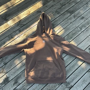 Brun hoodie från Adidas med huva - Säljer en brun Adidas hoodie i storlek S med klassisk vit logga på bröstet. Tröjan har huva med snörning, känguruficka och långa ärmar. Perfekt för chill dagar och streetstyle. Materialet är mjuk bomullsmix som känns skön mot huden.