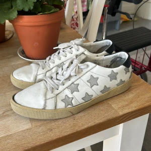 Saint Laurent sneakers med stjärnor - Coola vita sneakers från Saint Laurent med grå stjärnor på sidorna och svart detalj vid hälen. Skorna har snörning och platt sula, tillverkade i skinn. Perfekta för dig som vill sticka ut med en unik och trendig look. På vänster sko finns en liten defekt (se bilder), men som absolut går att laga bara genom lite textillim. Tillhöra låda följer tyvärr inte med