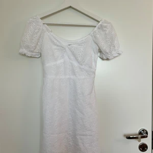 Vit kortklänning från Hollister XS - Superfin vit kortklänning från Hollister i broderad bomull med puffärmar och söt knutdetalj framtill. Klänningen har v-ringning, smockad rygg och romantiskt hålmönster. Perfekt för sommarens alla tillfällen!
