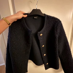 Svart kofta från Gina Tricot med guldknappar - Säljer en svart kofta från Gina Tricot med snyggt strukturstickat material och dekorativa guldknappar framtill. Koftan har rund halsringning och långa ärmar, perfekt för lager-på-lager och en stilren look.