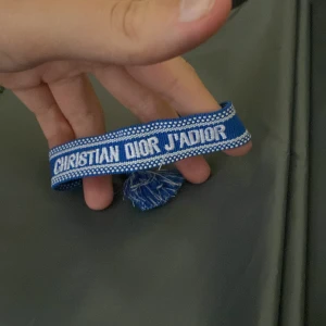 Blått Christian Dior J'Adior armband - Snyggt blått armband från Christian Dior med texten 'J'Adior' broderad i vitt. Armbandet har justerbara tofsar i blått och vitt och är gjort i ett mjukt, vävt tyg. Perfekt accessoar för att lyfta din outfit med en lyxig touch.