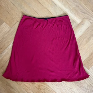 Mörkröd kortkjol med rosett - Enkel och snygg mörkröd kortkjol med liten svart rosett och tunn svart kant upptill. Kjolen har en lätt A-linjeform och är gjord i ett mjukt, tunt material som känns skönt mot huden. Perfekt för dig som gillar stilrena plagg med en söt detalj.