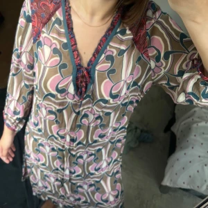 Mönstrad kortklänning med knyt - Säljer en kortklänning med färgglatt blommigt mönster i rosa, beige och blått. Klänningen har trekvartsärm, v-ringning med knyt och röda detaljer vid axlarna. Luftigt material och loose passform, perfekt för vår och sommar.