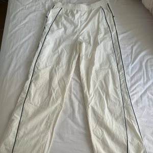 Vita tracksuit pants - Säljer ett par vita byxor med svarta ränder längs sidorna och elastisk midja. Byxorna har snörning vid benslut för justerbar passform. Perfekta för en chill och sportig look. Materialet känns lätt och bekvämt.