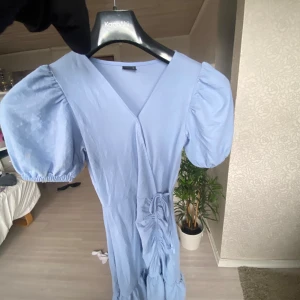 Ljusblå omlottklänning Gina Tricot - Supersöt ljusblå omlottklänning från Gina Tricot med puffiga korta ärmar och knyt i midjan. Klänningen har v-ringning och fina volangdetaljer framtill. Perfekt för dig som gillar en feminin och trendig stil.