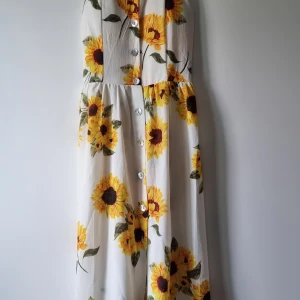 Vit klänning med solrosor och knappar - Långklänning med solrosmönster på vit botten. Klänningen har smala axelband, knappar framtill och en luftig, flowy passform. Perfekt för varma dagar när du vill sticka ut med färgglada blommor och somrig vibe.