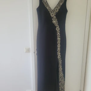 Svart långklänning med silverbroderi - Elegant svart långklänning med djup v-ringning och detaljerat silverbroderi längs urringningen och snett över framsidan. Ärmlös modell med figurnära passform och lång kjol, perfekt för bal eller fest. Broderiet ger en lyxig och unik känsla. Storlek 38/40