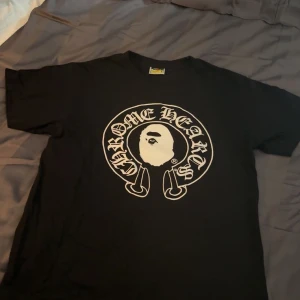 Svart BAPE x Chrome Hearts t-shirt S - Svart t-shirt från BAPE i samarbete med Chrome Hearts. Stor vit logga med ikoniskt ape-motiv framtill och Baby Milo-grafik samt Chrome Hearts-kors på ryggen. Klassisk passform och crewneck. Perfekt för dig som gillar streetwear och unika collabs.