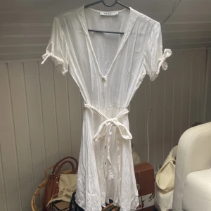 Vit kortklänning från NA-KD - Superfin vit kortklänning från NA-KD med v-ringning, knappar framtill och knytband i midjan. Perfekt för konfirmation/student. Använd en gång. 