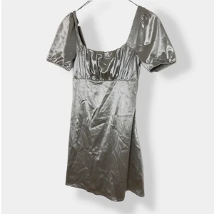 Silvrig kort klänning från H&M Divided - Supersnygg silvrig kort klänning från H&M Divided med glansigt satinliknande material. Klänningen har puffiga korta ärmar, fyrkantig ringning och lätt rynkade detaljer upptill. Perfekt för dig som vill sticka ut med en cool metallic vibe.