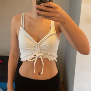 Vit croppad linnetopp  - Superfin vit croppad linnetopp från FB Sister med smala axelband och snygg snörning framtill. Går att skjutstera längden framtill med hjälp av snörena! Toppen har volangkant nertill och är perfekt för varma dagar. Mjuk och stretchig bomullskänsla som sitter skönt på kroppen.🤍🤍