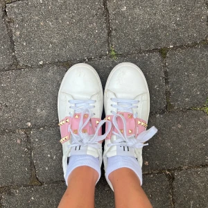 Valentino Rockstud sneakers vit/rosa- nästan helt nya - Snygga vita sneakers från Valentino med en bred rosa rem över vristen dekorerad med guldiga nitar. Skorna har klassisk snörning, rund tå och är tillverkade i skinn. Perfekt statement-piece för dig som gillar detaljer och lyxig streetstyle.