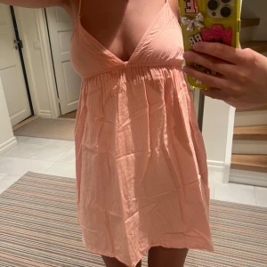 Ljusrosa kort sommarklänning - Supersöt ljusrosa kort klänning med tunna axelband och v-ringning. Klänningen har en flowy passform och är gjord i ett lätt bomullstyg med diskreta ränder. Perfekt för varma dagar och ger en chill vibe.