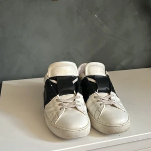Valentino Open sneakers vit/svart skinn - Snygga Valentino Open sneakers i vitt skinn med bred svart läderdetalj över mitten. Klassisk rund tå, platt sula och diskreta nitar längs hälen. Perfekta för dig som gillar stilrena och lyxiga sneakers med en twist.