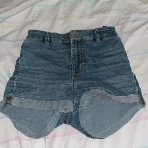 Blå jeansshorts från H&M Divided - Snygga blå jeansshorts från H&M Divided i klassisk denim med hög midja och uppvikta benslut. Shortsen har fem fickor, bälteshällor och dragkedja med knapp framtill. Perfekta för varma dagar och enkel att matcha med allt.
