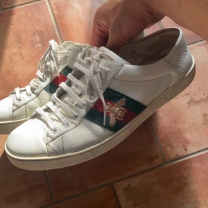 Gucci ace.  bi   🐝 ♦️ - Säljer ett par vita sneakers från Gucci i skinn med klassiska gröna och röda ränder på sidan samt en broderad guldfärgad bi. Skorna har snörning och platt sula. Perfekta för dig som vill sticka ut med en lyxig och ikonisk look.
