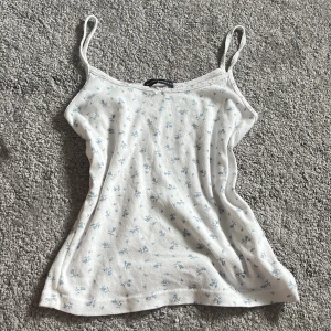 Vit blommig linnetopp Brandy Melville - Supersöt vit linnetopp från Brandy Melville med ljusblått blommönster och smala axelband. Toppen har en liten rosett framtill och är gjord i mjuk bomull. Köpt i London för ca 250 kr och är aldrig använd💗minsta acceptabla pris: 100kr