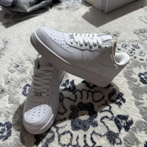 Nike Air Force 1 vita sneakers - Klassiska Nike Air Force 1 sneakers i helvitt skinn med perforerad tå, rund tå och platt sula. Snygg och tidlös modell med snörning och diskret Nike-logga på sidan. Perfekta för dig som gillar en clean och stilren look.