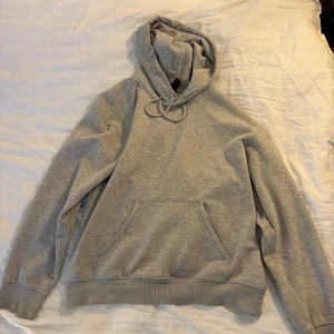 Grå hoodie  - En basic grå hoodie med huva och snörning, samt en stor magficka framtill. Tröjan har långa ärmar och ribbade muddar vid ärmslut och nederkant. Perfekt för chill dagar eller när du vill ha en enkel och stilren look.