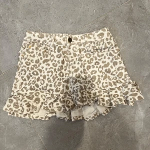 Leopardmönstrad kjol med volang - Snygga shorts med beige och brunt leopardmönster. Modellen har volangdetalj längst ner och klassisk femficksdesign. Stängs med knapp och dragkedja. Perfekta för dig som vill sticka ut lite extra i sommar.