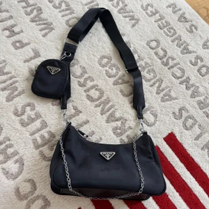 Svart Prada axelväska med kedja - Säljer en svart Prada axelväska i nylon med silverfärgade detaljer och kedja. Väskan har en justerbar axelrem och en liten avtagbar miniväska med dragkedja. Klassisk trekantslogga framtill och extra fack med dragkedja på baksidan.
