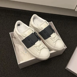 Valentino Open sneakers vit/svart skinn - Snygga Valentino Open sneakers i vitt skinn med bred svart rem över vristen och diskret logga på plösen. Klassisk rund tå, platt sula och coola nitar baktill. Perfekta för dig som gillar stilrena och lyxiga detaljer.