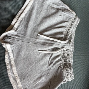 Grå mjuka shorts med snörning - Superbekväma grå shorts i mjukt bomullstyg med elastisk midja och snörning. De har vita kanter och en avslappnad passform, perfekta för chill dagar eller träning. Klassisk sportig look med kortare ben och rundade sidor.