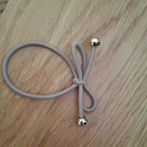 Beige armband med silverkulor - Stilrent beige armband i elastiskt material med två blanka silverfärgade kulor i ändarna. Armbandet har en enkel knutdetalj och passar perfekt för en clean och modern look.